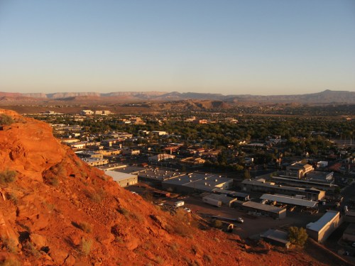 A panoramic view of a desert town with red rock formations and mountains in the background during sunset.