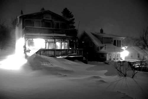 A snowy night scene featuring two houses, one illuminated, with deep snow covering the ground.