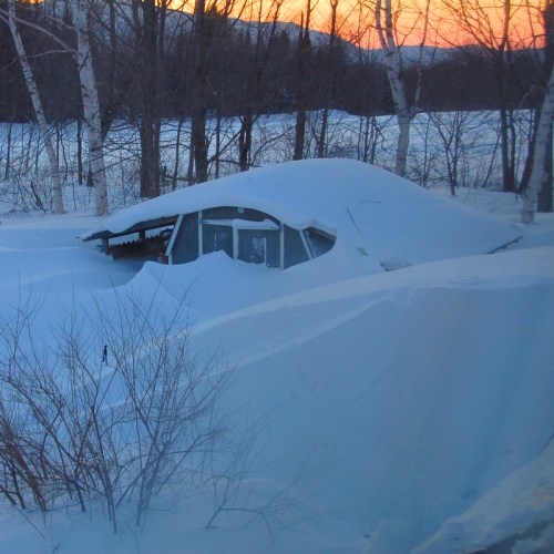 A snow-covered landscape at sunset, with a partially buried building and trees in the background.