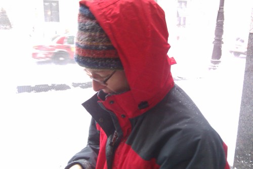 A person in a red jacket and colorful hat stands outside in heavy snow, looking down at their phone.