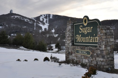 Sign for Sugar Mountain Skiing with snowy landscape and ski slopes in the background.