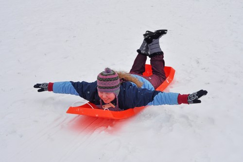 A child in winter clothing sleds down a snowy hill, arms outstretched, wearing a colorful hat and gloves.