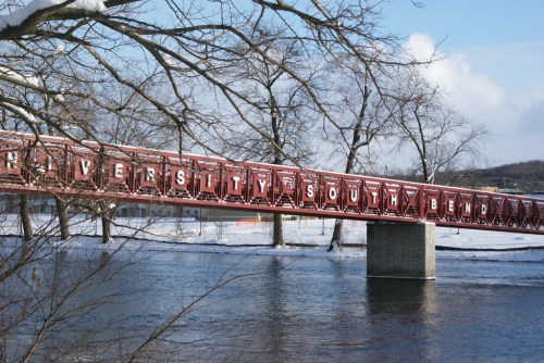A red pedestrian bridge over a river, surrounded by snow-covered trees and a clear blue sky.