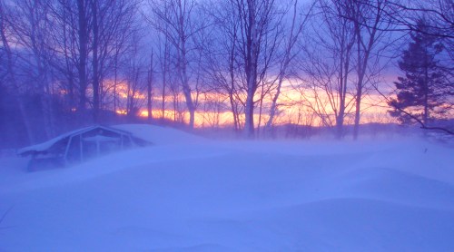 A snowy landscape at sunset, with trees silhouetted against a colorful sky and a cabin partially buried in snow.
