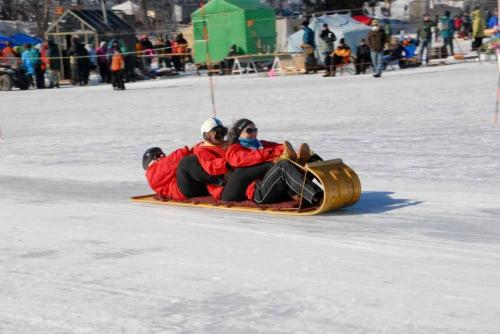 Three people in red jackets sledding down a snowy slope, surrounded by spectators and winter activities.