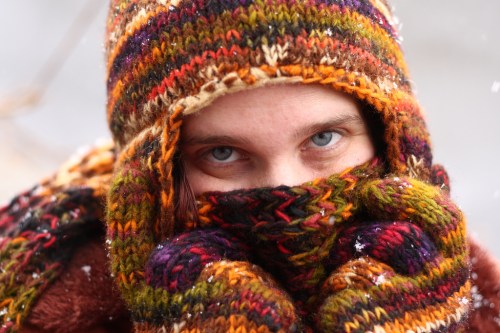 A person with bright blue eyes peeks out from a colorful knitted hat and scarf, surrounded by falling snow.