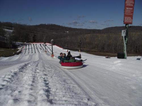 Two people enjoy tubing down a snowy slope on a sunny day, with a hill and trees in the background.