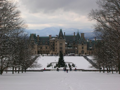 A grand mansion surrounded by snow-covered grounds and trees, with mountains in the background under a cloudy sky.
