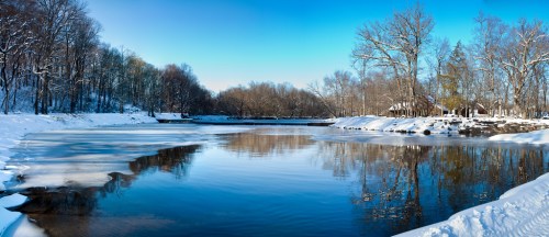 A serene winter landscape featuring a frozen river, snow-covered banks, and bare trees under a clear blue sky.
