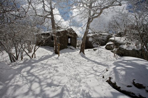 A snowy landscape with a stone building surrounded by trees and rocky terrain under a partly cloudy sky.