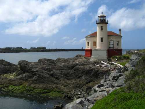 A red and white lighthouse stands on rocky shore, surrounded by water and green vegetation under a partly cloudy sky.