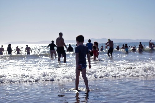 A child stands in shallow water at the beach, with people swimming in the background under a bright sky.