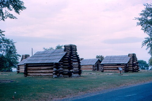 Historic log cabins set in a grassy area, with trees and a cloudy sky in the background.