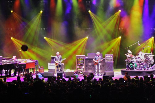 A live band performs on stage with colorful lights and a large audience in front, creating an energetic atmosphere.