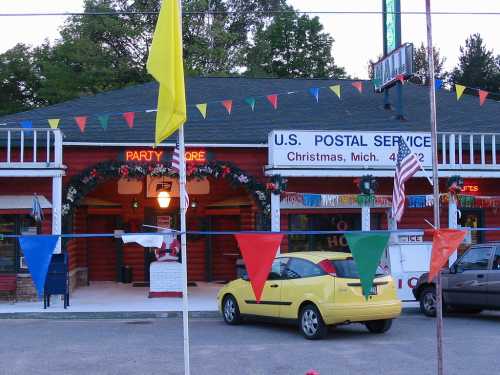 A yellow car parked in front of a festive U.S. Postal Service building in Christmas, Michigan, decorated with flags and lights.