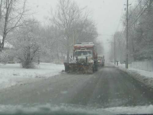 A snowplow clears a snowy road lined with trees, while snow falls heavily in the background.