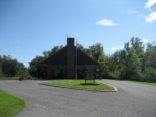 A small brick building with a sloped roof, surrounded by greenery and a clear blue sky. A sign is visible in front.