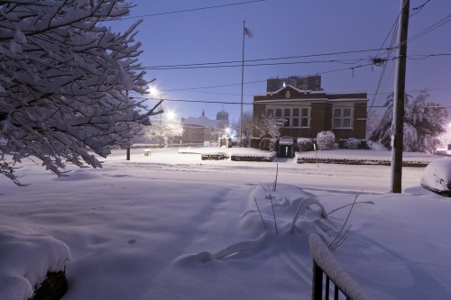 A snowy street scene at night, with a flag and a building in the background, illuminated by streetlights.