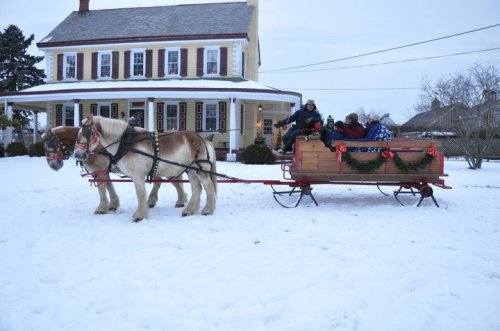 A horse-drawn sleigh carries passengers through snow in front of a large, festive house.
