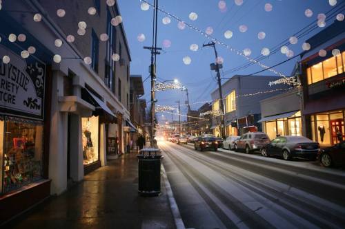 A street scene at dusk, lined with shops and festive lights, reflecting a cozy, holiday atmosphere.