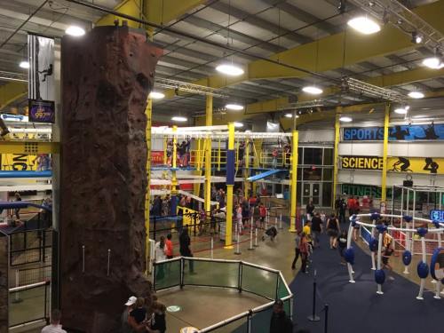 Indoor recreation center featuring a climbing wall, obstacle courses, and various activity areas filled with people.