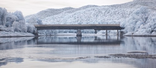 A serene landscape featuring a bridge over a calm river, surrounded by frosty trees and rolling hills.