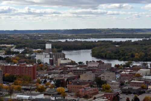 Aerial view of a riverside town with buildings, trees in autumn colors, and a river winding through the landscape.