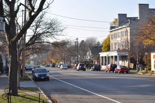 A quiet street lined with trees and parked cars, featuring a historic building and autumn foliage.
