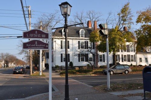 A street sign for S. Fifth St. near a white bed and breakfast building with autumn trees and a lamppost.