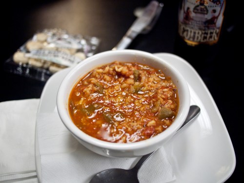 A bowl of hearty soup with vegetables, served on a white plate, alongside a packet of snacks and a drink.