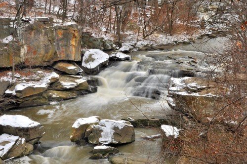A serene winter scene of a flowing river over rocky terrain, surrounded by bare trees and a dusting of snow.