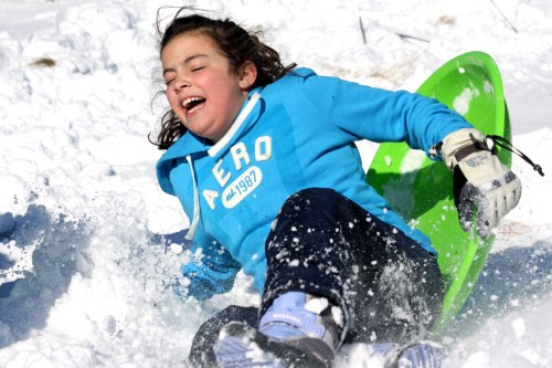 A child in a blue hoodie joyfully sleds down a snowy hill, laughing as snow sprays around them.