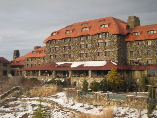 A large stone hotel with a red roof, surrounded by snow and greenery, set against a cloudy sky.