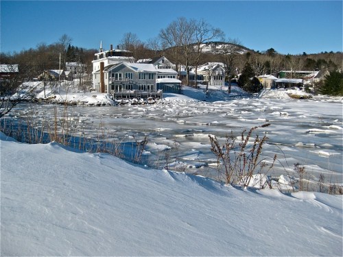 Snow-covered landscape with a frozen river and houses in the background, under a clear blue sky.