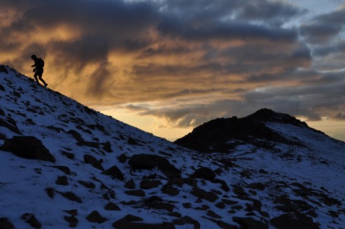 A silhouetted figure climbs a snowy hillside against a dramatic sunset sky with clouds.