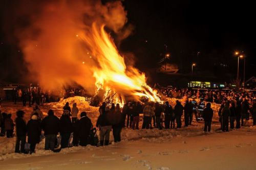 A large bonfire illuminates a snowy night as a crowd gathers around, enjoying the warmth and spectacle.