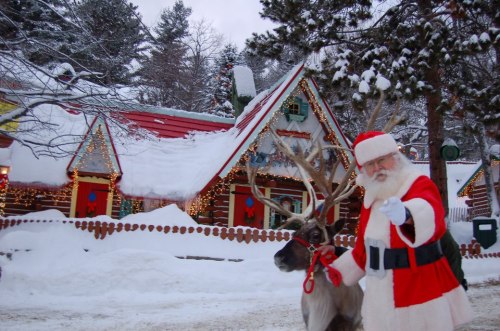 Santa Claus in a red suit waves beside a reindeer in a snowy village with festive decorations.
