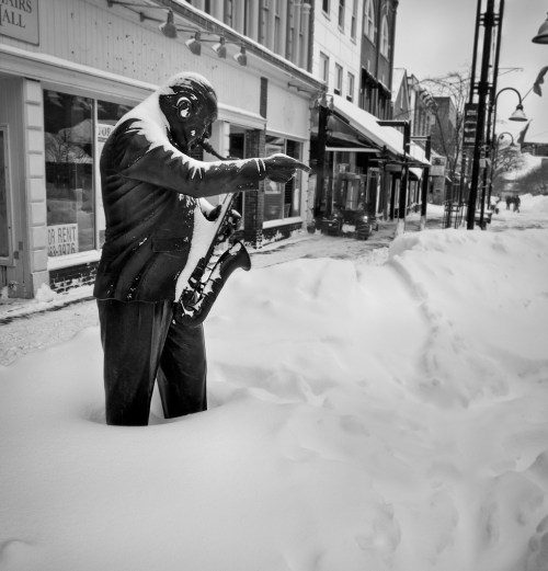 A snow-covered street scene featuring a statue of a man playing the saxophone, surrounded by deep snow and empty storefronts.