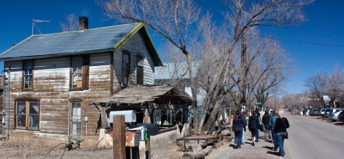 A group of people walks along a street lined with bare trees and an old, weathered wooden house.