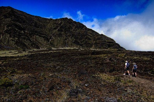 Two hikers walking on a rocky trail with a mountainous landscape and blue sky in the background.