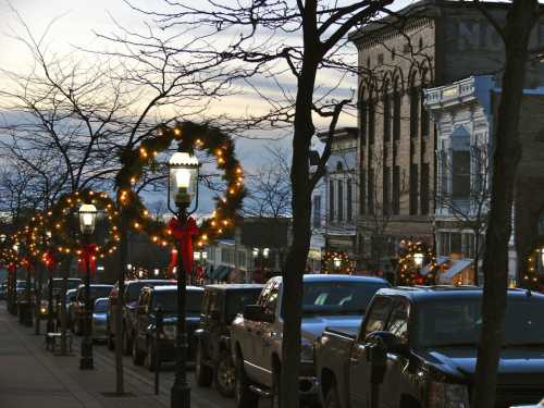 A festive street lined with decorated trees and wreaths, illuminated by lights, with parked cars and historic buildings.