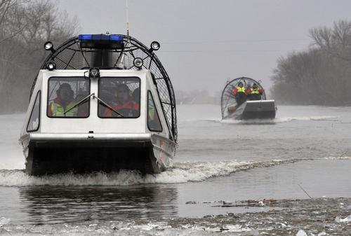 Two airboats navigate through a flooded area, with crew members visible in bright safety gear.