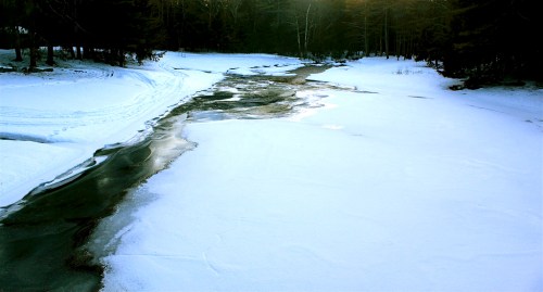 A serene winter landscape featuring a partially frozen river winding through a snowy field and trees.