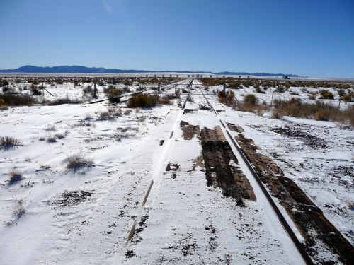 Abandoned railway tracks stretch through a snowy landscape under a clear blue sky, with distant mountains on the horizon.