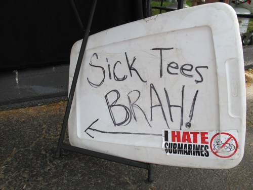 A sign on a white board reads "SICK TEES BRAH!" with an arrow pointing left and a sticker saying "I HATE SUBMARINES."