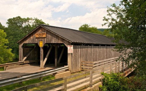 A wooden covered bridge with a metal roof, surrounded by trees and a clear sky, featuring a walkway and warning signs.
