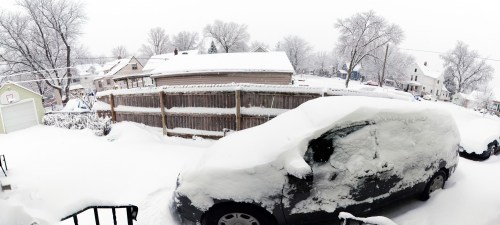 A snowy scene showing a car covered in snow, with houses and trees in the background under a gray sky.