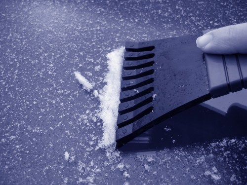 A hand using a black ice scraper to remove snow from a car's surface. The image has a blue tint.