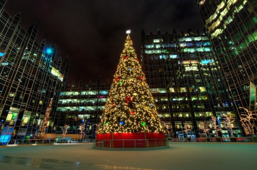 A brightly lit Christmas tree adorned with decorations stands in a plaza surrounded by modern glass buildings at night.
