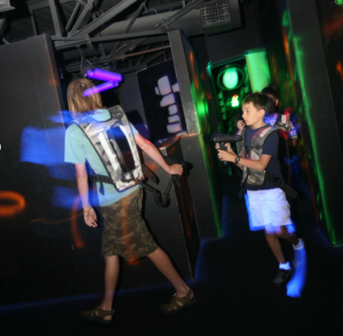 Two kids in a dark laser tag arena, wearing vests and holding laser guns, with colorful lights in the background.
